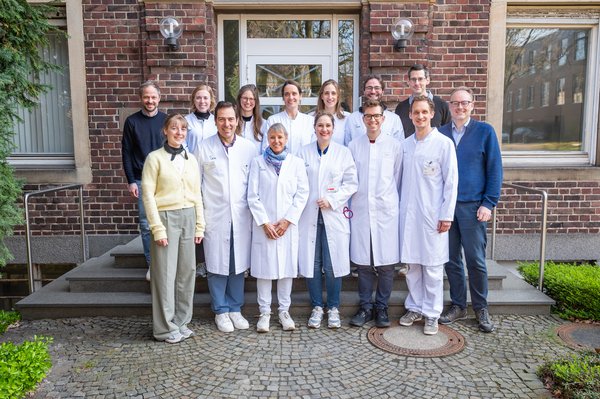 UKM | Interdisziplinäres Team mit 14 Personen. Sie stehen vor einem Klinkerbau in zwei Reihen und lachen; die meisten tragen einen weißen Kittel
