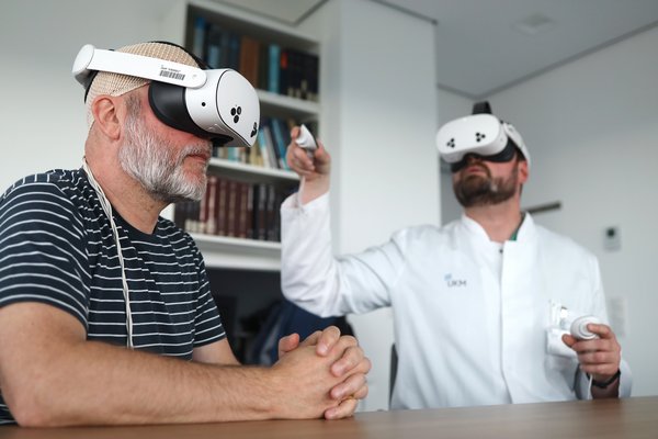UKM | Patient und Neurochirurg sitzen am Tisch und haben weiße VR-Brillen auf. Der Arzt hält Controller in jeder Hand.