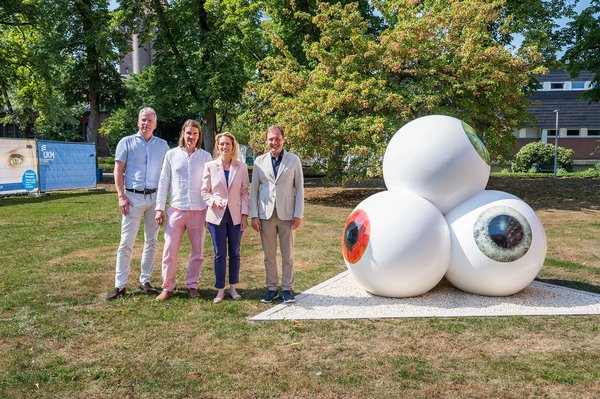 Auf einer Rasenfläche: Eine Skulpur mit drei großen weißen Kugeln, auf denen jeweils eine Pupille und Iris in Braun, Grau, Grün zu sehen ist und die in verschiedene Richtungen gerichtet sind; die vierte Kugel mit blauer Iris ist nicht zu sehen. Links daneben stehen der Kaufmännische Direktor, der Künstler, die Direktorin der Augenklink und der Ärztliche Direktor des UKM