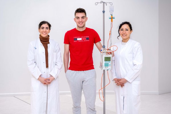 UKM | Zwei Ärztinnen im weißen Kittel stehen neben einem Lupus-Patienten im roten T-Shirt, der sich an einem Infusionsständer hält.