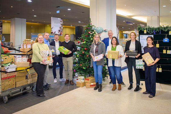 UKM | Neun Personen halten eingepackte Weihnachtsgeschenke in den Händen. Sie stehen vor dem geschmückten Weihnachtsbaum im Eingangsbereich der UKM-Cafeteria, weitere Päckchen stapeln sich an der Seite.