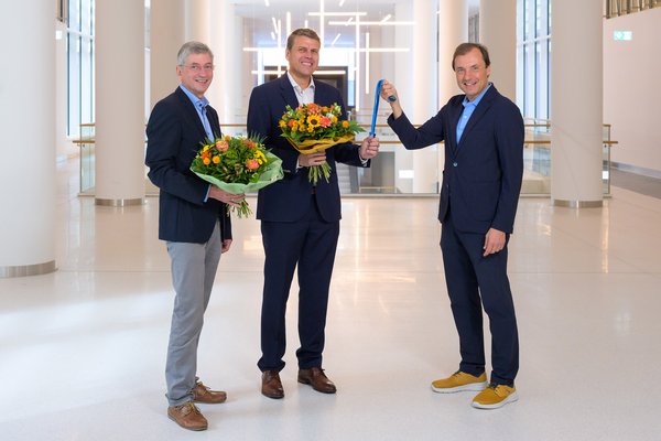 Drei Männer im Anzug schauen lachend in die Kamera, zwei haben einen Blumenstrauß in der Hand