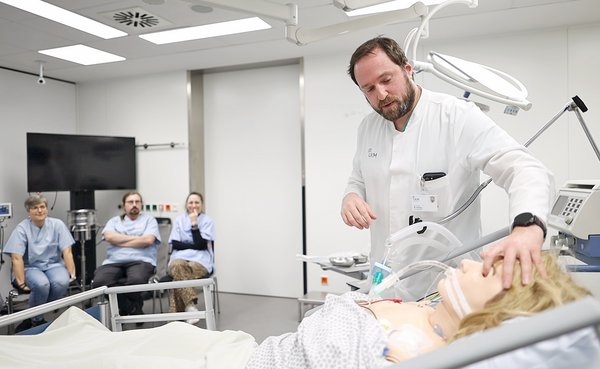 UKM | Ein Arzt fasst mit einer Hand einer Übungspuppe an die Augen. Drei Mitarbeitende sitzen gegenüber und schauen zu.