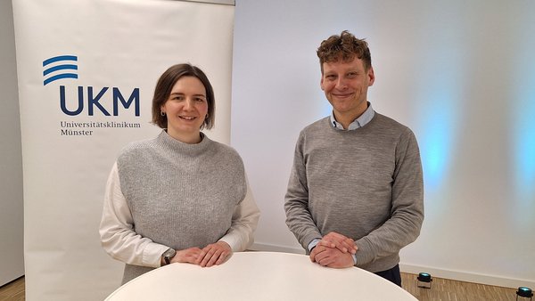 UKM | Pflegerin und Arzt an einem weißen Stehtisch. Im Hintergrund ein weißes Rollup mit dem UKM-Logo