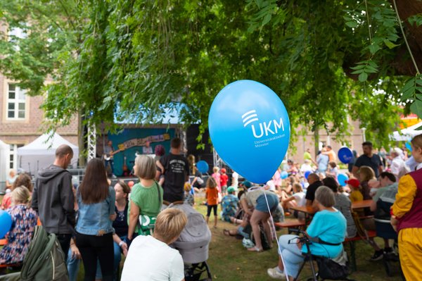 Blauer Luftballon mit UKM-Logo, viele Leute stehend und sitzend unter Bäumen