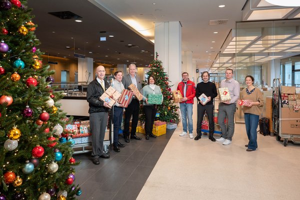 UKM | Fünf Mitarbeitende des UKM und drei Mitarbeitende der Hilfsorganisationen stehen vor einem geschmückten Weihnachtsbaum und Rollwagen mit Päckchen. Sie halten verpackte Geschenke in den Händen und lachen. Im Vordergrund ein weiterer Weihnachtsbaum mit bunten Kugeln und kleinen Lämpchen