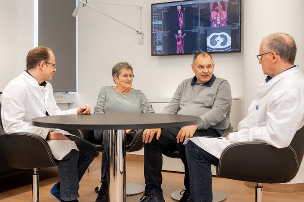 UKM | Knochenmarktransplantationszentrum | Zwei Ärzte sowie Patient und Ehefrau sitzen an einem runden Tisch. Monitor mit Bildern aus bildgebenden Verfahren an der Wand