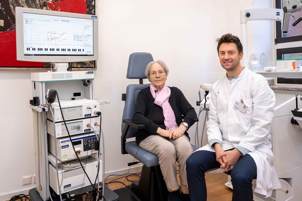 UKM | Behandlungszimmer mit Geräten und Monitor in der Phoniatrie. Patientin und Arzt und sitzen nebeneinander und schauen in die Kamera.
