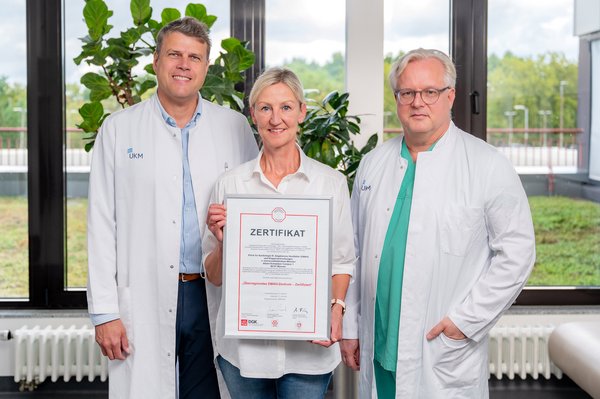 UKM | Presse | Zwei Ärzte im weißen Kittel, dazwischen hält die Sekretärin das eingerahmte Zertifikat