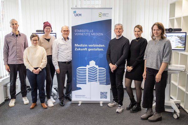 UKM | Sieben Mitarbeiterinnen und Mitarbeiter der Stabsstelle Vernetzte Medizin. Sie stehen in einem Büro links und rechts neben einem hochen Banner mit der Aufschrift Stabsstelle Vernetzte Medizin, Medizin verbinden, Zukunft gestalten