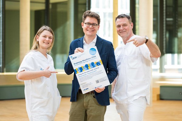UKM | Zwei Pflegekräfte aus der Onkologie und der WTZ-Geschäftsführer, der ein Plakat in den Händen hält. Alle lächeln.