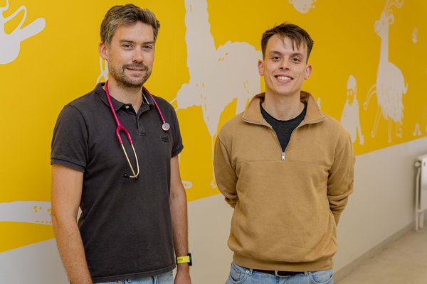 UKM | Zwei Männer vor einer Wand mit weißen-gelben Motiven. Der eine trägt ein braunes Poloshirt und hat ein Stethoskop um den Hals, der andere trägt ein beiges Sweatshirt.