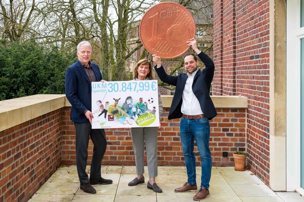 UKM | Zwei Männer und eine Frau auf einer Terrasse. Sie halten ein Plakat mit der Spendensumme in den Händen und einen großen Cent in die Höhe.